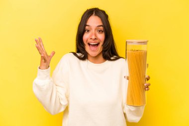 Sarı arka planda izole edilmiş spagetti kavanozunu tutan genç İspanyol kadın hoş bir sürpriz alıyor, heyecanlı ve el kaldırıyor..