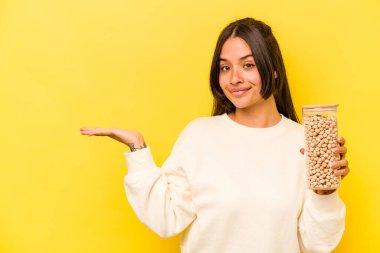 Sarı arka planda izole edilmiş bir nohut kavanozu tutan genç İspanyol kadın bir avuç içinde fotokopi alanı ve belinde bir el daha olduğunu gösteriyor..