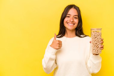 Sarı arka planda izole edilmiş nohut kavanozunu tutan genç İspanyol kadın gülümsüyor ve başparmağını kaldırıyor.