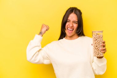 Sarı arka planda izole edilmiş nohut kavanozunu tutan genç İspanyol kadın zaferden sonra yumruğunu kaldırıyor, kazanan konsept..