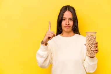 Sarı arka planda izole edilmiş nohut kavanozu tutan genç İspanyol kadın parmak ile bir numarayı gösteriyor..