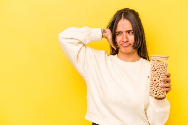 Genç İspanyol kadın elinde bir nohut kavanozu tutuyor sarı arka planda izole edilmiş kafasının arkasına dokunuyor, düşünüyor ve bir seçim yapıyor..