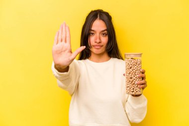 Genç İspanyol kadın elinde bir nohut kavanozu tutuyordu. Sarı arka planda izole bir şekilde duruyordu. Ellerini uzatmış, dur işareti göstererek seni engelliyordu..