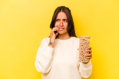 Elinde nohut kavanozu tutan genç İspanyol kadın sarı arka planda izole edilmiş tırnak yiyor, gergin ve endişeli..