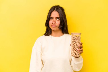 Sarı arka planda izole edilmiş nohut kavanozunu tutan genç İspanyol kadın şaşkın ve kararsız hissediyor..