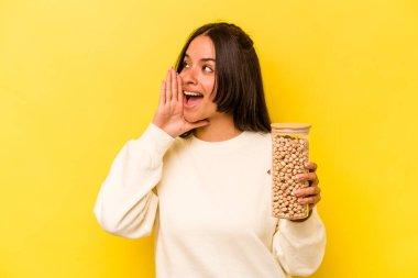 Sarı arka planda izole edilmiş nohut kavanozunu tutan genç İspanyol kadın bağırıyor ve ağzı açık bir şekilde avuç içi tutuyor..