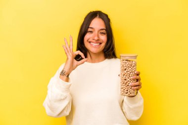 Elinde nohut kavanozu tutan genç İspanyol kadın sarı arka planda izole edilmiş neşeli ve kendinden emin bir şekilde iyi niyet gösteriyor..