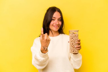 Sarı arka planda izole edilmiş nohut kavanozunu tutan genç İspanyol kadın sanki seni davet ediyormuş gibi işaret ediyor..