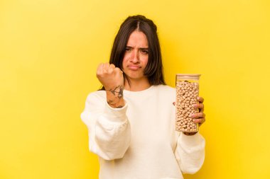 Sarı arka planda izole edilmiş nohut kavanozu tutan genç İspanyol kadın kameraya yumruk gösteriyor, agresif yüz ifadesi..