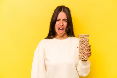 Sarı arka planda izole edilmiş nohut kavanozunu tutan genç İspanyol kadın çok sinirli ve agresif çığlıklar atıyor..