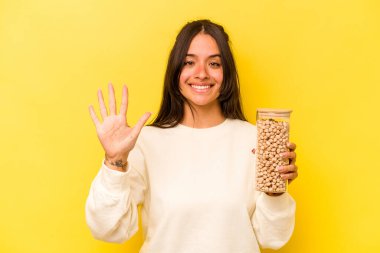 Sarı arka planda izole edilmiş nohut kavanozunu tutan genç İspanyol kadın gülümseyerek beş numarayı parmaklarıyla gösteriyor..