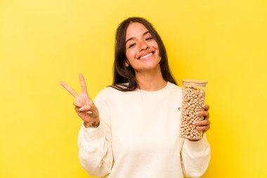 Sarı arka planda izole edilmiş bir nohut kavanozu tutan genç İspanyol kadın neşeli ve kaygısız parmaklarıyla barış sembolü gösteriyor..