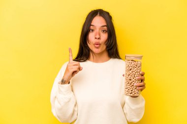 Sarı arka planda izole edilmiş bir nohut kavanozu tutan genç İspanyol kadının harika bir fikri var, yaratıcılık kavramı..