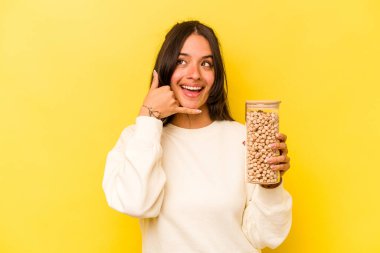 Sarı arka planda izole edilmiş nohut kavanozu tutan genç İspanyol kadın parmaklarıyla bir cep telefonu hareketi gösteriyor..