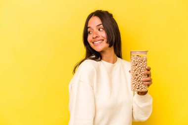 Sarı arka planda izole edilmiş nohut kavanozunu tutan genç İspanyol kadın gülümsüyor ve keyifli görünüyor..