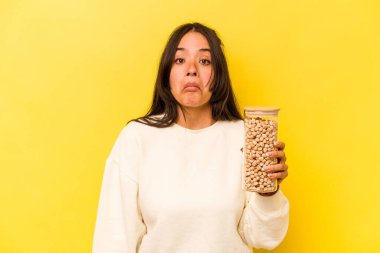 Sarı arka planda izole edilmiş nohut kavanozunu tutan genç İspanyol kadın omuzlarını silker ve gözleri açık kalır..