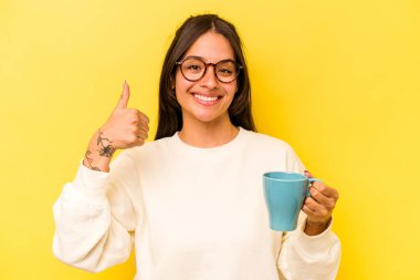 Sarı arka planda izole edilmiş fincanı tutan genç İspanyol kadın gülümsüyor ve başparmağını kaldırıyor.