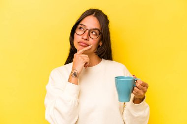 Sarı arka planda bir kupa tutan genç İspanyol kadın şüpheli ve şüpheci bir ifadeyle yana bakıyor..