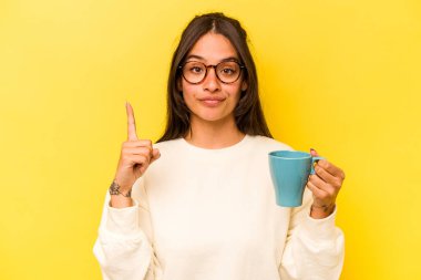 Sarı arka planda elinde fincanla gezen Latin kökenli genç bir kadın bir numarayı parmaklarıyla gösteriyor..