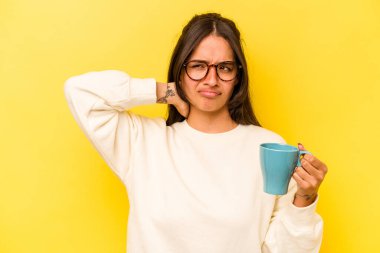 Sarı arka planda izole edilmiş bir kupa tutan genç İspanyol kadın kafasının arkasına dokunuyor, düşünüyor ve bir seçim yapıyor..