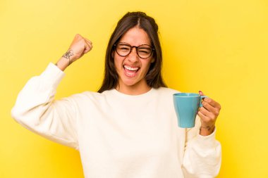 Sarı arka planda izole edilmiş fincanı tutan genç İspanyol kadın zaferden sonra yumruğunu kaldırıyor, kazanan konsept..