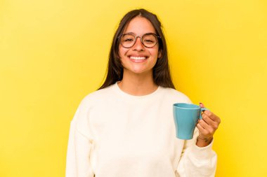 Sarı arka planda saklanmış bir kupa tutan genç İspanyol kadın mutlu, güler yüzlü ve neşeli..