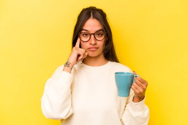 Sarı arka planda izole edilmiş bir kupa tutan genç İspanyol kadın parmağıyla tapınağı işaret ediyor, düşünüyor ve bir göreve odaklanıyor..