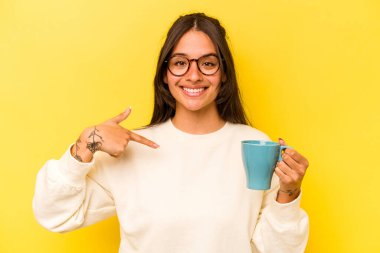 Sarı arka planda izole edilmiş bir kupa tutan genç İspanyol kadın gömlek fotokopi alanını işaret ediyor, gururlu ve kendinden emin.