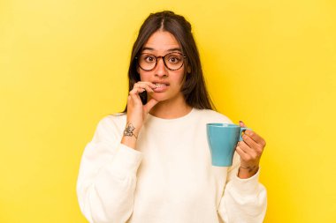 Sarı arka planda izole edilmiş, tırnak yiyen, gergin ve endişeli bir kupa tutan genç İspanyol kadın..
