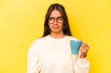 Sarı arka planda izole edilmiş bir kupayı tutan genç İspanyol kadın şaşkın ve kararsız hissediyor..