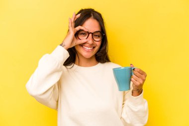 Sarı arka planda izole edilmiş bir kupa tutan genç İspanyol kadın heyecanlı..
