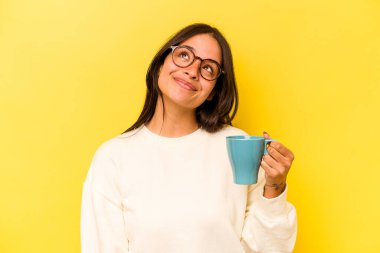 Genç İspanyol kadın sarı arka planda izole edilmiş bir kupa tutuyor. Amaçlarına ve amaçlarına ulaşma hayali kuruyor.