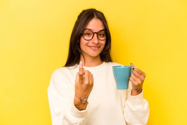 Sarı arka planda izole edilmiş bir kupa tutan genç İspanyol kadın sanki seni davet ediyormuş gibi seni işaret ediyor..