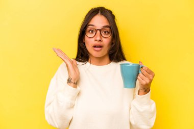 Sarı arka planda bir kupa tutan genç İspanyol kadın şaşırdı ve şok oldu..