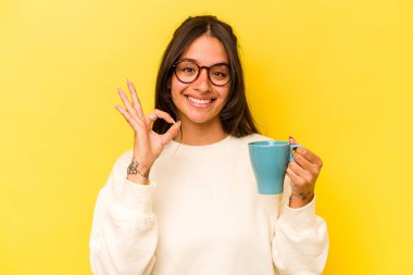 Sarı arka planda izole edilmiş bir kupa tutan genç İspanyol kadın neşeli ve kendinden emin..