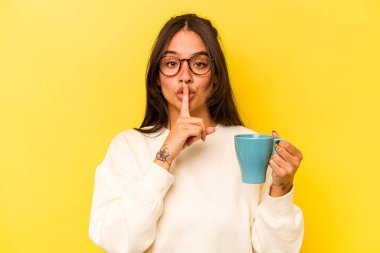 Sarı arka planda bir kupa tutan genç İspanyol kadın ya sır saklıyor ya da sessizlik istiyor..