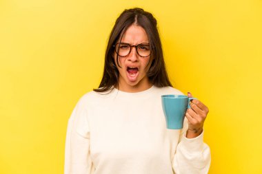 Sarı arka planda izole edilmiş bir kupa tutan genç İspanyol kadın çok sinirli ve agresif bağırıyor..