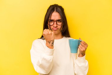 Sarı arka planda izole edilmiş bir kupa tutan genç İspanyol kadın kameraya yumruğunu gösteriyor, agresif yüz ifadesi..