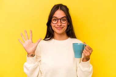 Sarı arka planda izole edilmiş bir kupa tutan genç İspanyol kadın gülümseyerek beş numarayı parmaklarıyla gösteriyor..