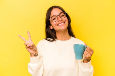 Sarı arka planda izole edilmiş bir kupa tutan genç İspanyol kadın neşeli ve kaygısız parmaklarıyla barış sembolü gösteriyor..