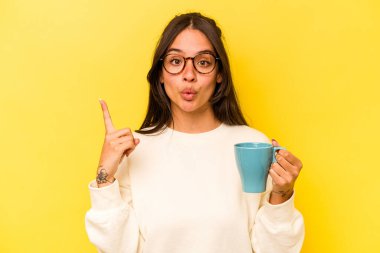 Sarı arka planda izole edilmiş bir kupa tutan genç İspanyol kadının harika bir fikri var, yaratıcılık kavramı..