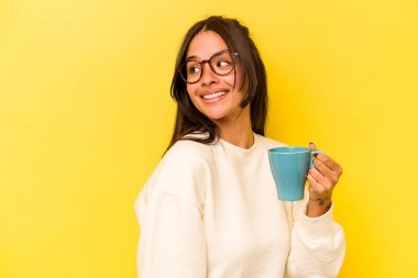 Sarı arka planda saklanmış bir kupa tutan genç İspanyol kadın gülümsüyor, neşeli ve keyifli görünüyor..