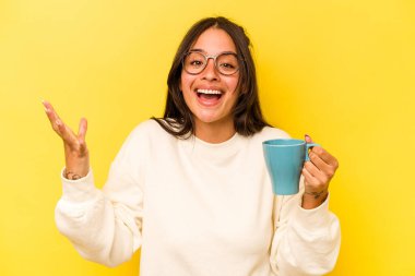 Sarı arka planda bir kupa tutan genç İspanyol kadın hoş bir sürpriz alıyor, heyecanlı ve el kaldırıyor..