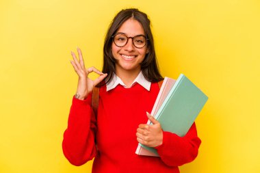 Genç İspanyol öğrenci, sarı arka planda izole edilmiş neşeli ve kendinden emin, iyi bir jest yapıyor..