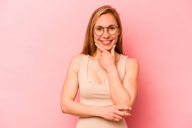 Pembe arka planda izole edilmiş genç beyaz kadın gülümsüyor ve kendinden emin, elleriyle çenesine dokunuyor..
