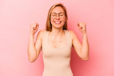 Pembe arka planda izole edilmiş genç beyaz kadın zaferi, tutkuyu ve coşkuyu kutluyor, mutlu bir ifade..