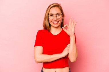 Pembe arka planda izole edilmiş genç beyaz kadın bir göz kırpıyor ve elinde iyi bir el hareketi tutuyor..