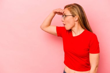 Pembe arka planda izole edilmiş genç beyaz kadın uzaklara bakıyor ve elini alnına koyuyor..