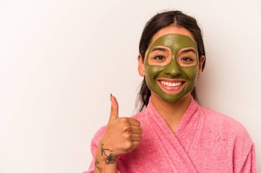 Beyaz arka planda soyutlanmış yüz maskesi takan genç İspanyol kadın gülümsüyor ve başparmağını kaldırıyor.