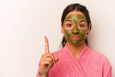 Yüz maskesi takan genç İspanyol kadın beyaz arka planda bir numarayı parmağıyla gösteriyor..